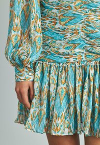Vestido multicolorido con patrones abstractos en turquesa y naranja, mangas largas abullonadas, falda fruncida y detalles de botones en los puños.