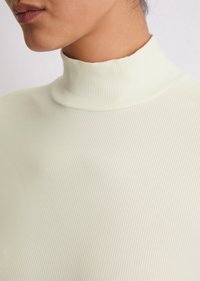 Ribattu turtleneck toppi vaaleassa kerma niin, että se sopii istuvuudeltaan ja hienovaraiselta tekstuuriltaan, jossa on korkea pääntie ja sileä viimeistely.