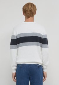 Persona bionda che indossa un maglione bianco con righe orizzontali grigie e nere, abbinato a jeans blu, in piedi contro uno sfondo neutro.