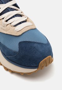 Blue sneaker featuring a combination of mesh and suede materials, with beige accents and white laces; textured rubber outsole visible.