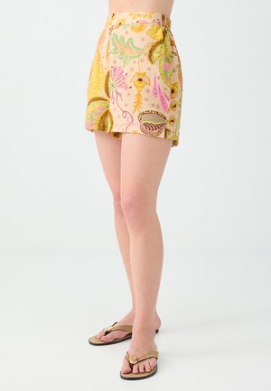 Personne portant un short jaune à motif floral et des sandales tongs beige, debout sur un fond blanc.