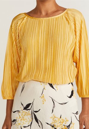 Žena nosí žlutou plisovanou blůzu s tříčtvrtečními rukávy v kombinaci s bílou sukní s černými a žlutými květinovými vzory.