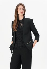 Blazer nero con vestibilità sartoriale, caratterizzato da un design delle spalle strutturato, tasche frontali con zip e arricchito da un top in pizzo nero sottostante.
