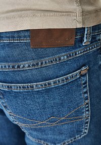 Dark blue denim jeans featuring a leather label with embossed text. The back pockets display a distinct stitching pattern.