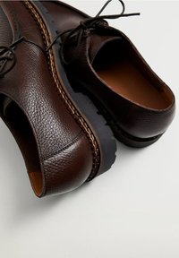 Chaussures à lacets en cuir marron avec finition texturée et semelles épaisses noires, placées sur une surface gris clair.