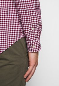 Red and white gingham check shirt with a buttoned cuff, paired with olive green pants. Fabric appears smooth with a crisp pattern.