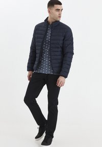 Marineblå pufferjacka med horisontella sömmar, en ståkrage, över en mönstrad grå skjorta och svarta byxor, i kombination med mörka sneakers.