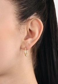 Pendiente de aro dorado con una superficie texturizada, forma circular y pequeño acento. Llevado en el lóbulo, destacando su diseño contra la piel.