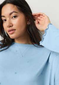 Lyseblå sweater med et tekstureret stjernemønster, ribbet halsudskæring og flæsekant ved ærmerne. Blødt stof med en afslappet pasform.