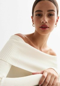 Off-shoulder, geribd gebreid top in off-white. Kenmerkt zich door een brede halslijn en een gestructureerde stof, met handen die op de stof rusten.