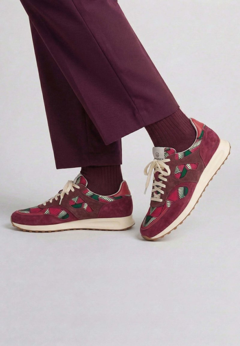 Personne portant un pantalon bordeaux et des chaussettes assorties avec des baskets bordeaux au motif coloré, debout sur un sol clair.