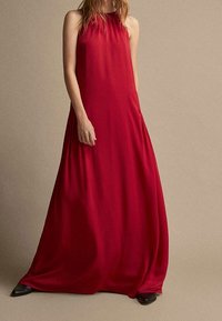 Robe longue rouge en tissu lisse, avec un design sans manches, un col montant et une silhouette fluide. Accessoirisée avec des chaussures noires.