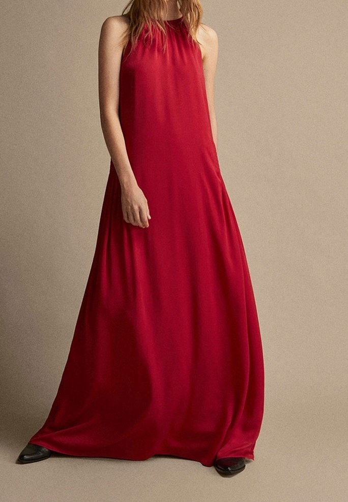 Robe longue rouge en tissu lisse, avec un design sans manches, un col montant et une silhouette fluide. Accessoirisée avec des chaussures noires.