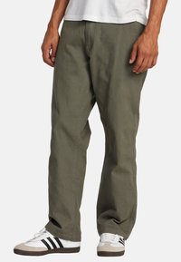 Green cotton pants with a relaxed fit and straight leg. Features visible stitching and side pockets. Worn with white and black sneakers.