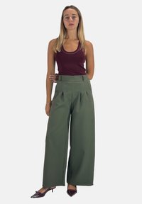 Groene geplooide high-waisted broeken gemaakt van een gladde stof, gecombineerd met een bordeauxrood tanktop en donkere puntige hakken.