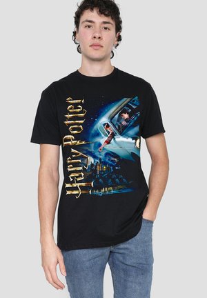Junger Mann trägt ein schwarzes Harry-Potter-T-Shirt mit fliegendem Auto und Hogwarts-Schloss, kombiniert mit blauen Jeans, steht vor schlichtem Hintergrund.