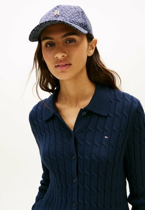 Frau mit langen dunklen Haaren trägt eine blaue, strukturierte, geknöpfte Strickjacke und eine gemusterte blaue Baseballkappe mit einem Logo auf der Vorderseite.