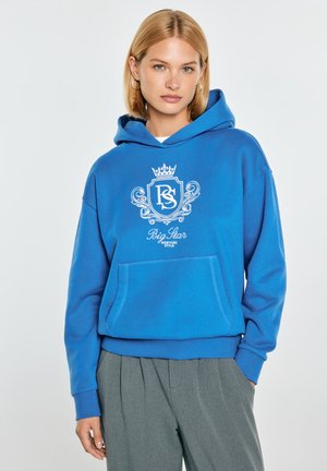 Młoda kobieta w jaskrawo niebieskiej bluzie z kapturem z białym herbem i tekstem "Big Star New York Style", w połączeniu z szarymi plisowanymi spodniami.