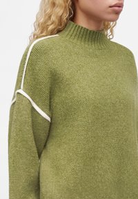 Personne portant un pull en maille vert olive avec un passepoil blanc sur l'épaule et le haut du bras, montrant le cou et une partie du visage de profil.