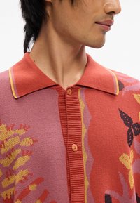 Korteermet strikket polo skjorte i varme rosa nyanser. Har en strukturert krage, to oransje knapper og et blomstertrykk i gult og rødt.