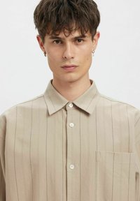 Beige knappeskjorte med vertikale striper, korte ermer og brystlomme; laget av lett stoff med en lett teksturert overflate.