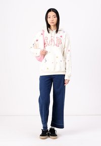 Sudadera crema con patrones florales y logo "GAP" en rojo, combinada con jeans de pierna ancha de color azul oscuro y zapatillas negras con suelas beige.