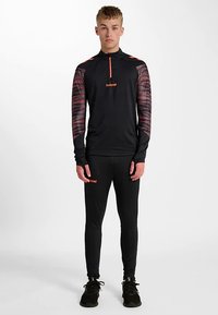 Svart atletisk topp med långa ärmar och krage med halvdragkedja, med orange accenter och ett mönstrat mönster på ärmarna; kombinerad med svarta leggings.