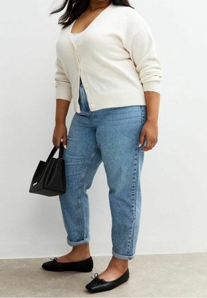 Kobieta w kremowym swetrze na guziki, jasnoniebieskich podwiniętych dżinsach, czarnych balerinach, trzymająca czarną torebkę na jednolitym tle.