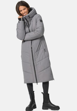Grå vadderad lång kappa med hög krage och framsidans dragkedja. Har vinklad söm och sidofickor. Bärs med svarta klumpiga stövlar.