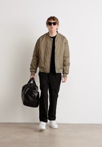 Veste bomber vert olive, chemise noire, pantalon noir, baskets blanches, tenant un sac de sport noir, lunettes de soleil. Design minimaliste, texture douce.