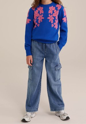 Giovane che indossa un maglione blu con motivi floreali rosa, pantaloni cargo in denim a gamba larga e sneakers bianche, in piedi con le mani in tasca.