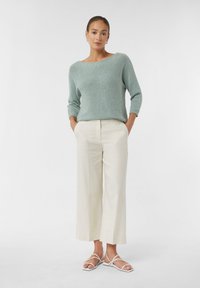 Hellblauer, gestrickter Pullover mit weit geschnittenem Ausschnitt, kombiniert mit cremefarbenen, weit geschnittenen Hosen und weißen, flachen Sandalen mit Riemchen. Durchgehend glatte Textur.