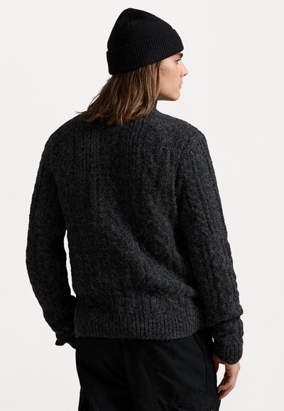 Persoon die een zwarte gebreide beanie en een donkergroene gebreide trui draagt, met de rug naar de camera gekeerd, tegen een eenvoudige witte achtergrond.