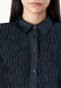 Černá plisovaná blůza s hladkým límečkem, předním zapínáním na knoflíky a texturovanou tkaninou s vertikálními pruhy a jemným leskem.
