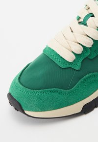 Green sneaker featuring a mix of smooth synthetic and textured suede materials, cream laces, black sole, and stitched detailing.