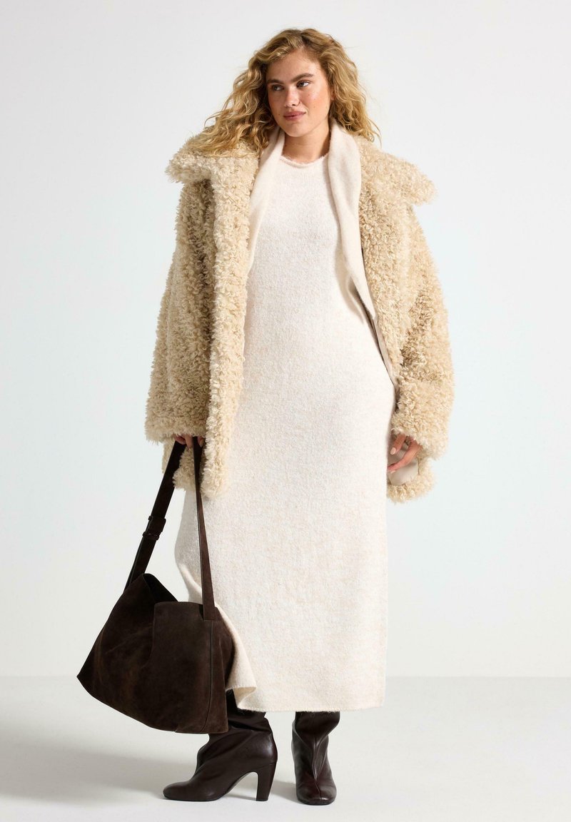 Kvinde iført en beige fuzzy frakke over en lang cremefarvet kjole, holder en stor mørkebrun suede taske, og har mørkebrun hæl støvler på.