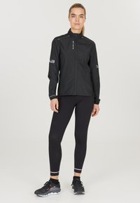 Schwarze leichte Jacke mit hohem Kragen, durchgehendem Reißverschluss und reflektierenden Details; kombiniert mit schwarzen Leggings und Laufschuhen mit Akzenten.