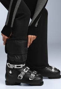 Fekete síbakancsok fémlakatokkal, párhuzamosan párnázott fekete nadrágcsatokkal, melyek gyűrött textúrával és kontrasztos mintával rendelkeznek az ujjakkal.