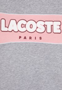 Camiseta de algodón gris claro con una banda horizontal rosa llamativa que lleva el texto "LACOSTE" en blanco y "PARIS" en rojo oscuro debajo.