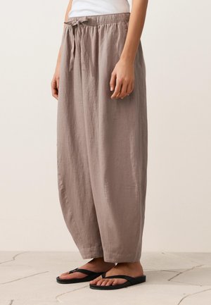 Relaxed-fit taupe linen pants with elastic waist and drawstring, worn with black flip-flops and a white top, standing on a light floor.