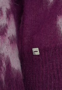 Tissu tricoté violet avec une texture douce et duveteuse, avec une étiquette en argent gravée "Mandla Grace" attachée au vêtement.