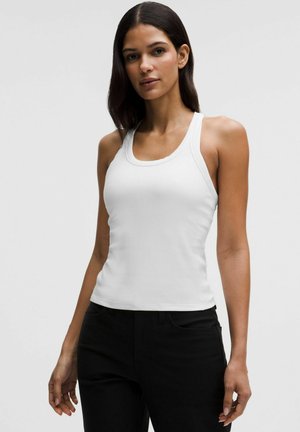 Femme aux longs cheveux bruns portant un débardeur blanc sans manches et un pantalon noir, se tenant devant un arrière-plan gris clair uni.