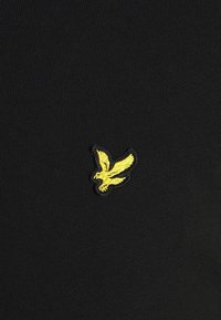 Musta neulottu kangas, jossa vasemmalla puolella pieni keltainen kirjailtu lintulogo. Pintatekstuuri on sileä ja istuva.