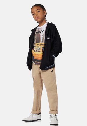 Zwarte zip-up jack met witte accenten, grafisch t-shirt met een stadsafbeelding in gele tinten, beige broek en witte schoenen met zwarte zolen.