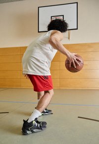 Sorte basketball sko med grønne detaljer, røde shorts og en hvit trøye, som dribler en basketball på et gymgulv. Basketballkurv synlig.