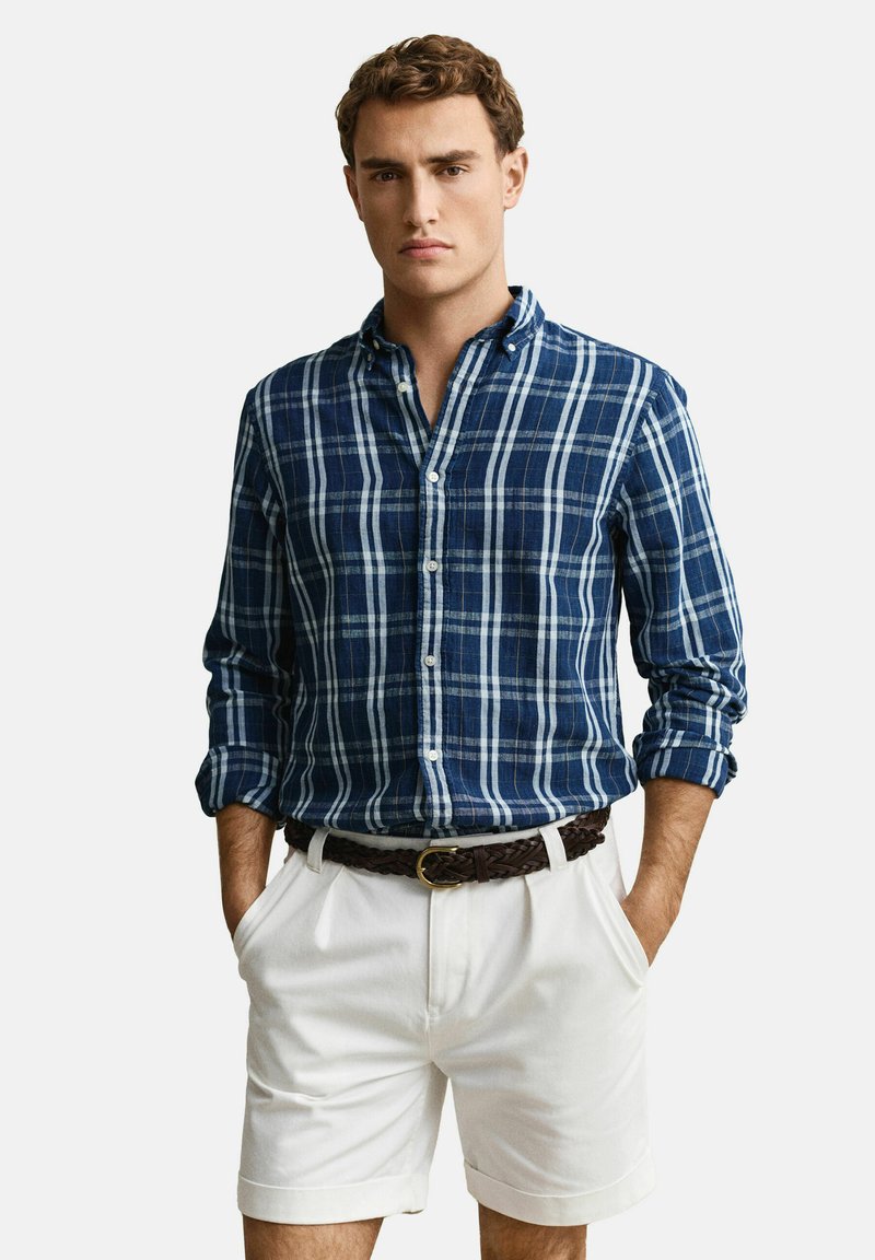 Giovane uomo che indossa una camicia blu a quadri con maniche arrotolate, pantaloncini bianchi e una cintura intrecciata marrone, in piedi con le mani nelle tasche.