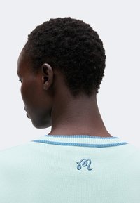 Personne aux cheveux courts, bouclés et foncés, portant un haut bleu clair avec un motif brodé bleu dans le dos près de l'encolure.