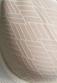 Béžová podprsenka s texturovaným, geometrickým síťovaným designem. Tkanina má vrstvu malých, protínajících se linií vytvářejících mřížkový vzor.