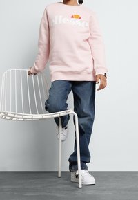 Sudadera rosa con el logo "ellesse", corte relajado. Jeans de mezclilla azul, zapatillas blancas. Persona apoyando la mano en una silla de alambre blanca, de pie sobre un pie.