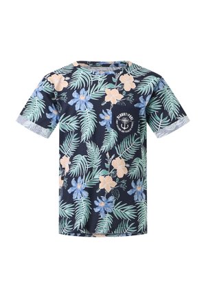 Dreng iført marineblå T-shirt med blomsterprint, blå jeans, hvide sneakers, holder gule solbriller og kigger til siden mod en ensfarvet baggrund.